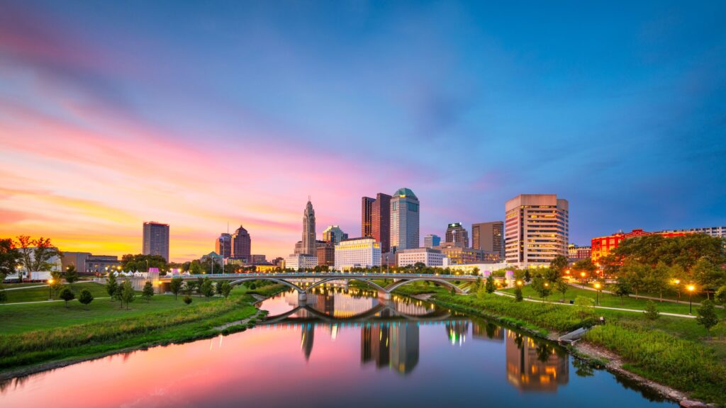 Central Columbus skyline along the Scioto River with LeVeque Tower, Rhodes State Office Tower, modern glass high-rises, and arched bridges at dusk.